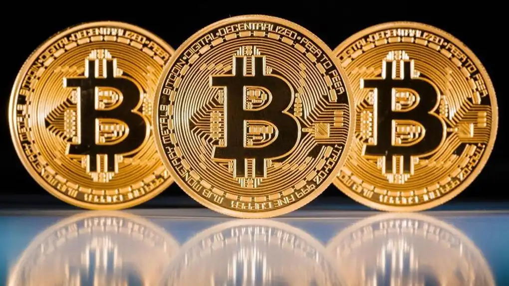 Three symbolic Bitcoin coins standing in a row with their reflection showing on the surface in front of them.